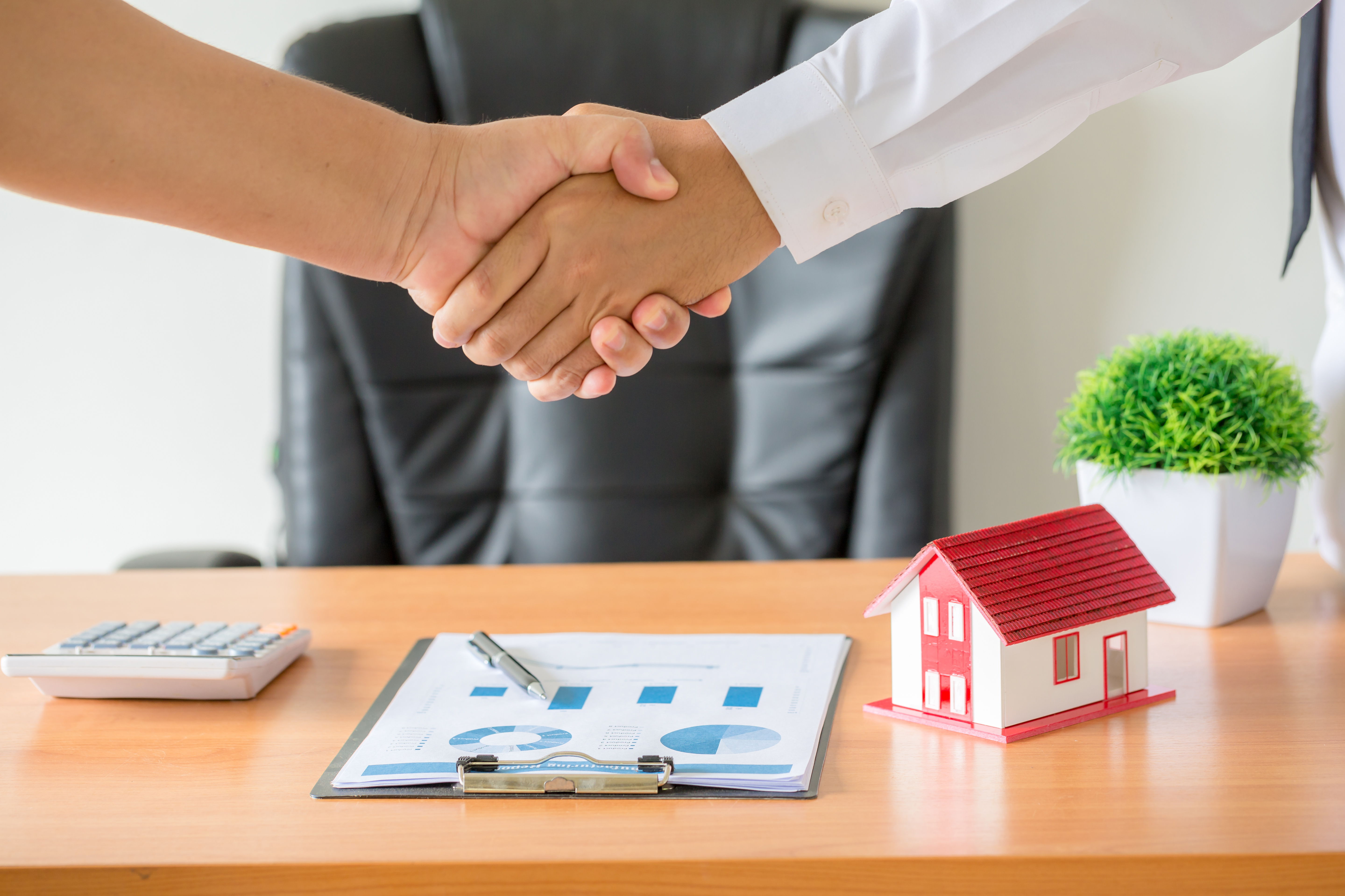 Handshake over a desk with a model house, documents, and calculator, symbolizing a real estate transaction decision.