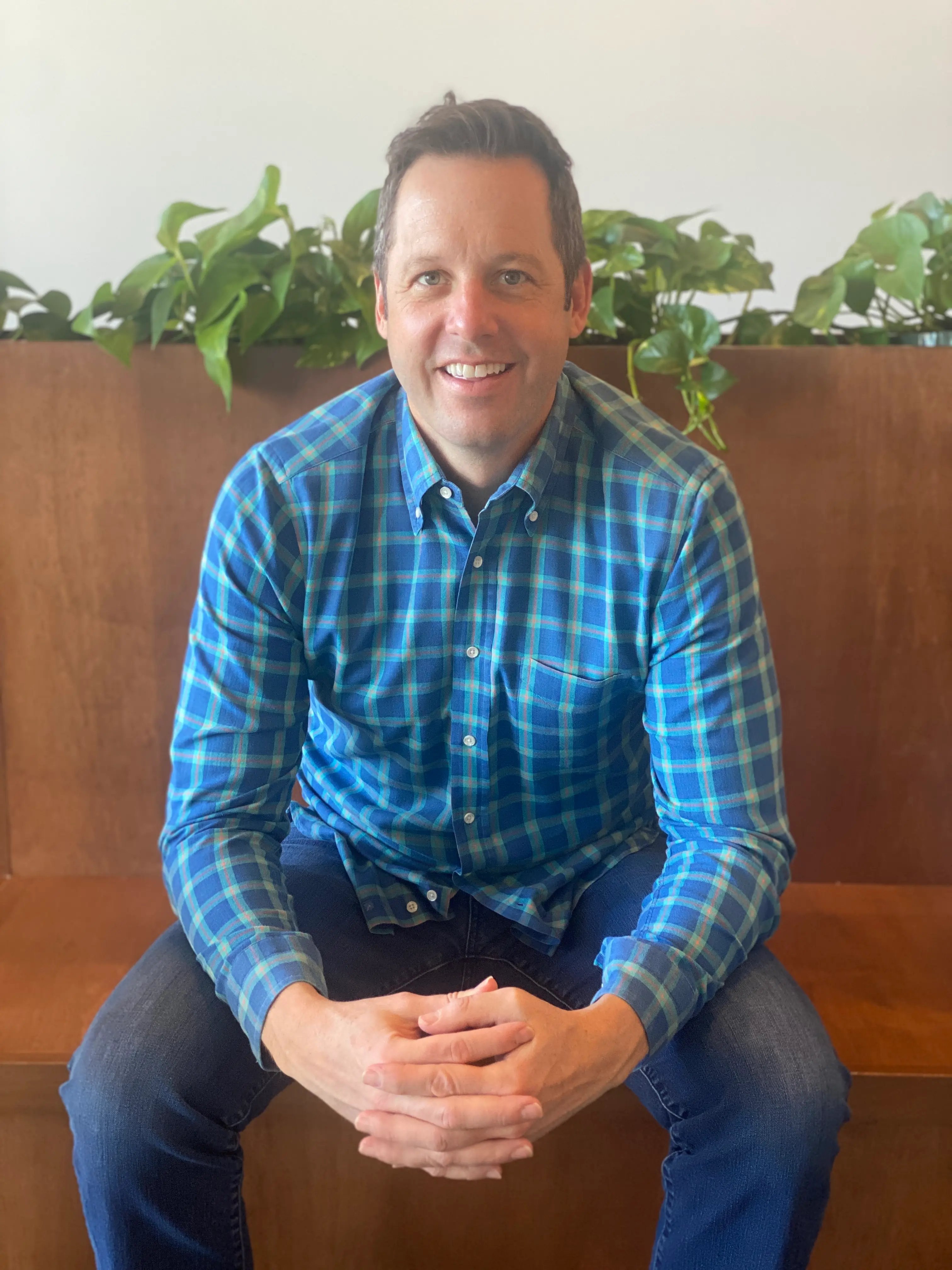 A man in a blue plaid shirt seated indoors, with plants in the background. Related to choosing between cash home buyers or hiring a realtor.