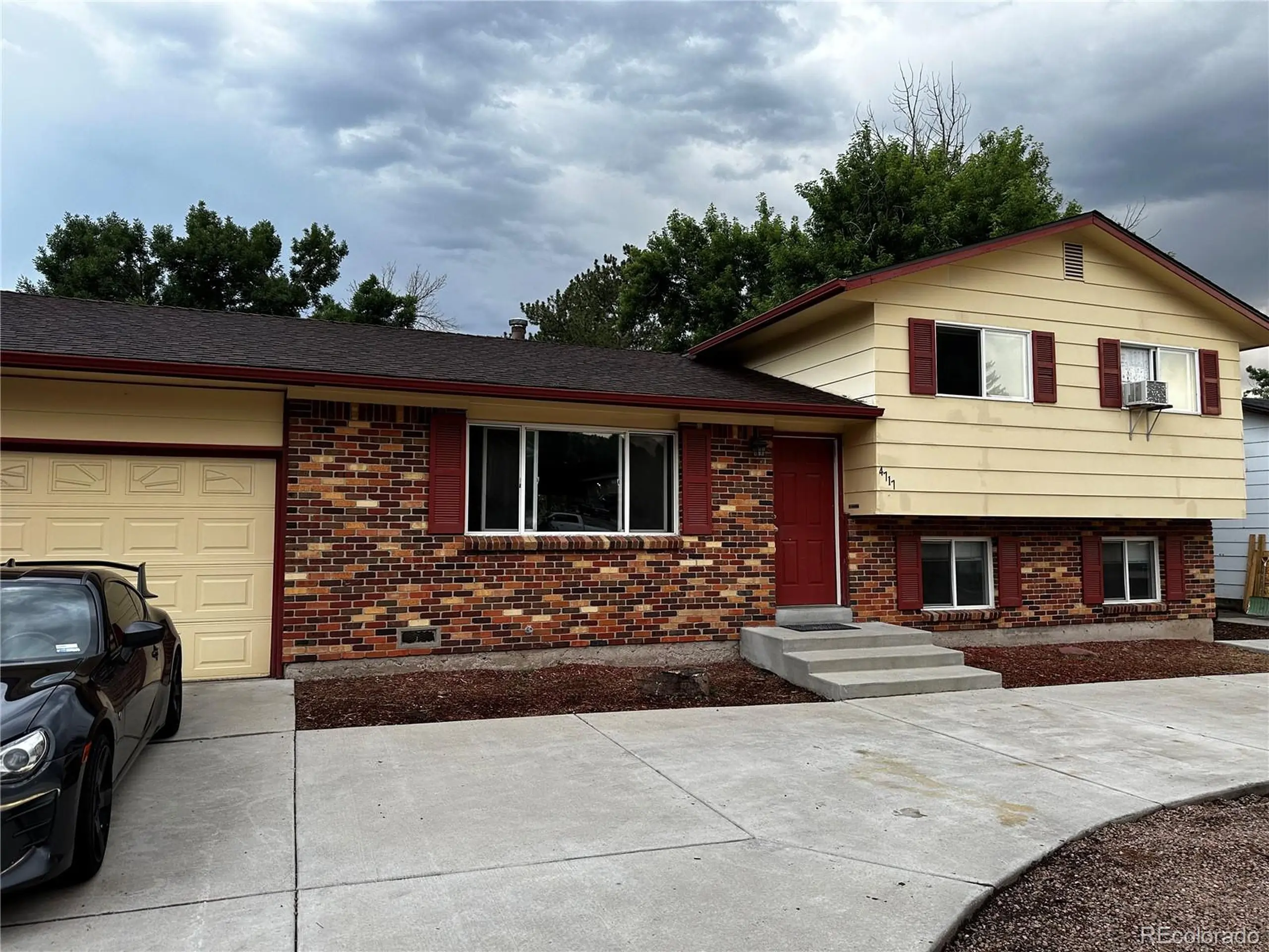 Exterior of a split-level home with a beige and brick facade, featuring a tidy driveway and a parked car, under an overcast sky.