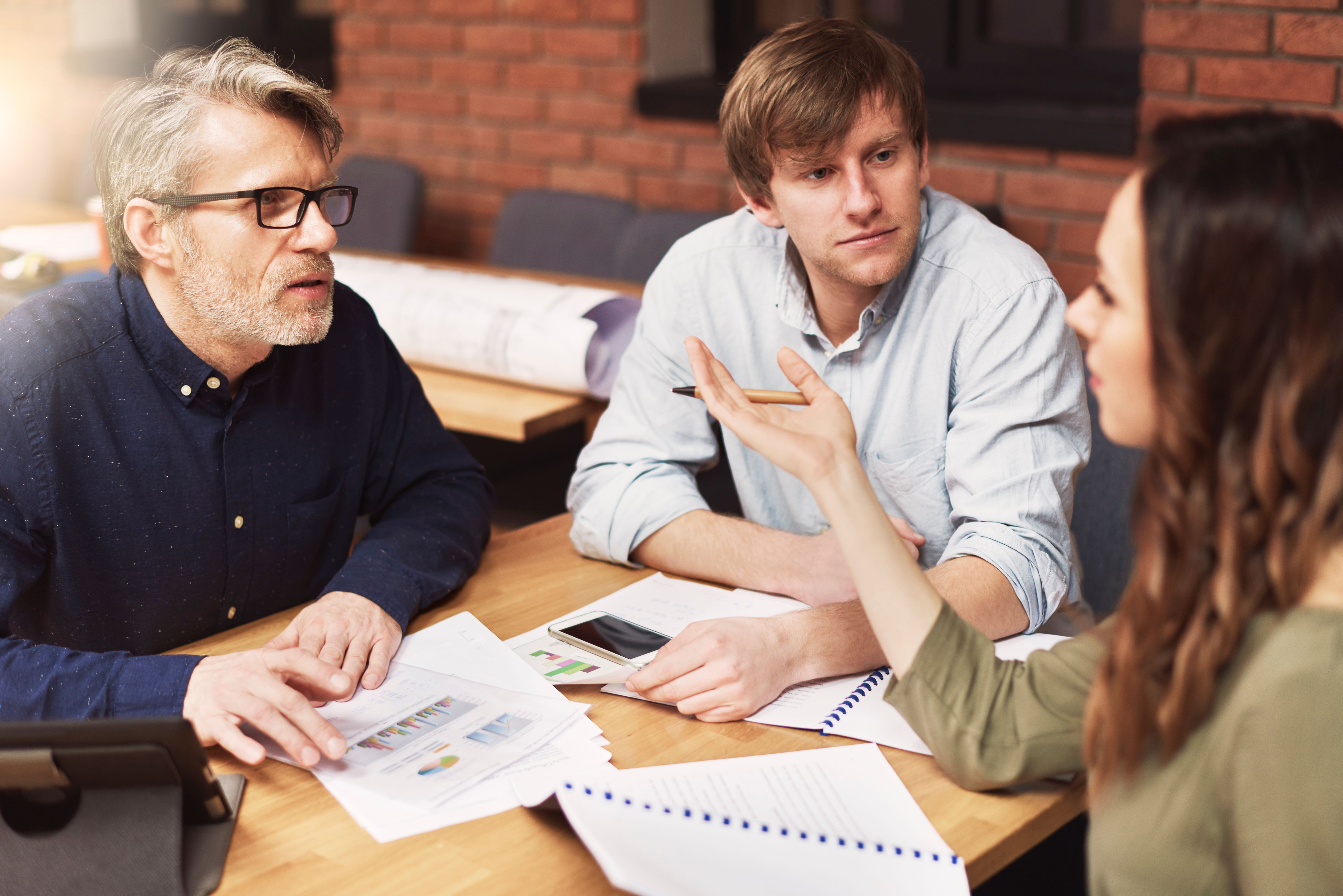 Three people discussing plans at a table with charts and documents, reflecting strategies for selling old homes profitably.