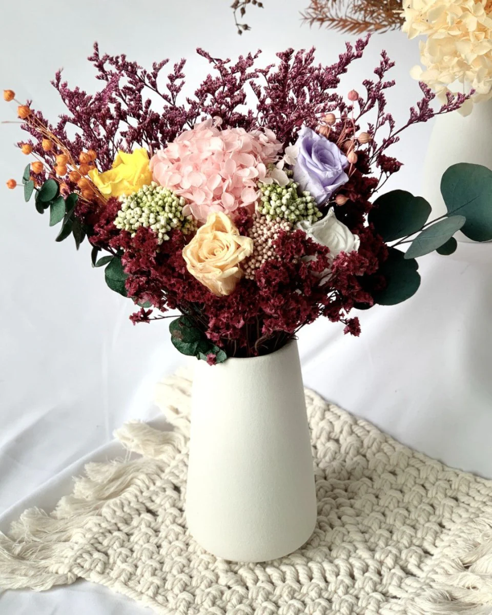 A white vase with preserved flowers, including roses and hydrangeas, on a textured cream mat, showcasing an elegant home decor idea.