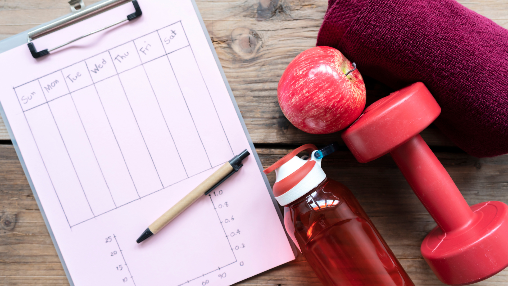 Workout plan clipboard, apple, towel, water bottle, and dumbbell on wooden surface.