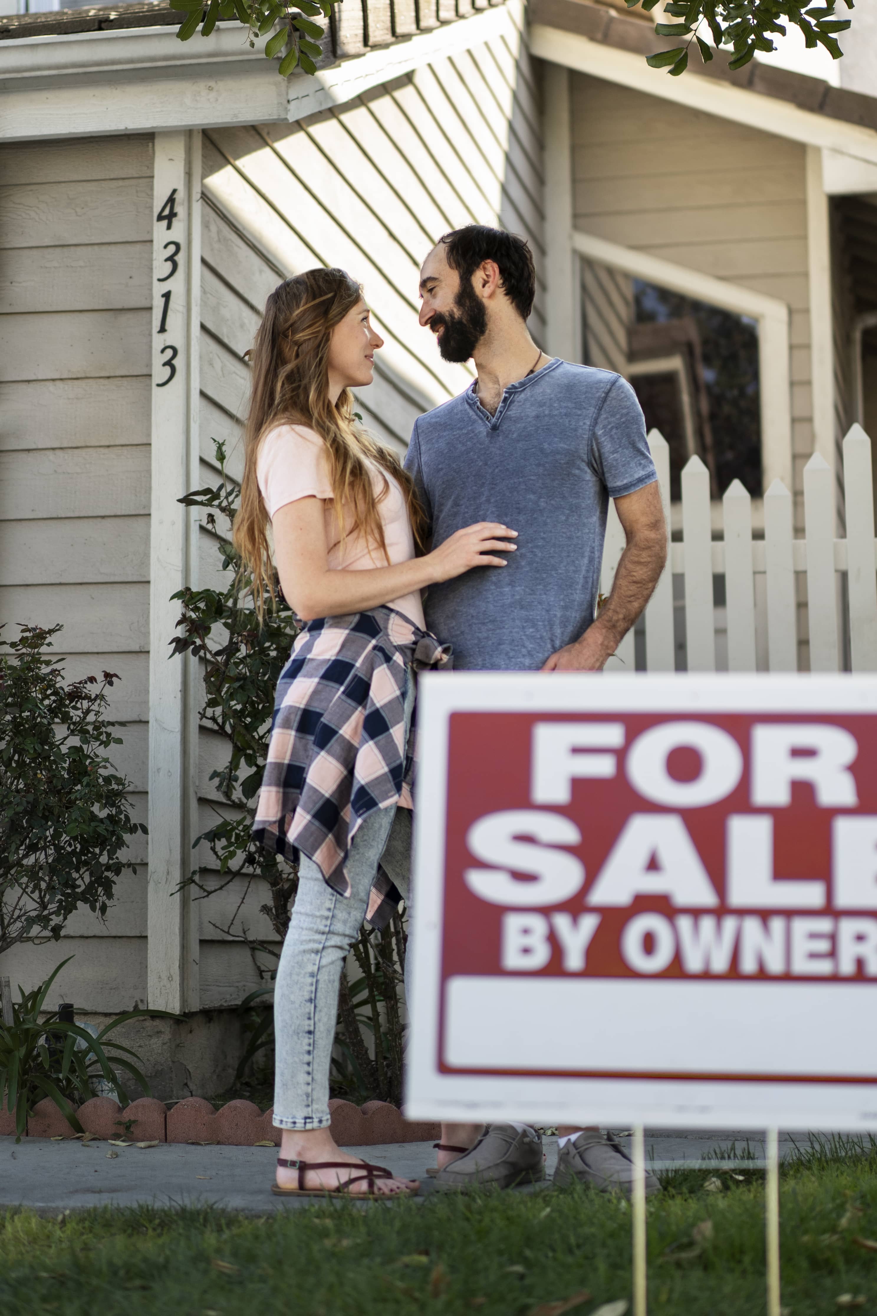 Couple stands outside their house with a “For Sale By Owner” sign, highlighting a DIY option for selling homes quickly.
