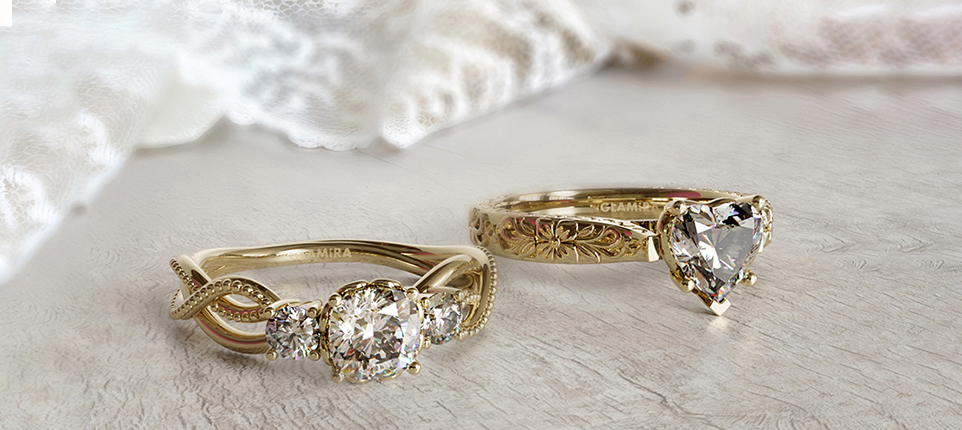 Two gold engagement rings: one with a round diamond and intricate band, the other with a heart-shaped diamond and floral details, on a lace background.