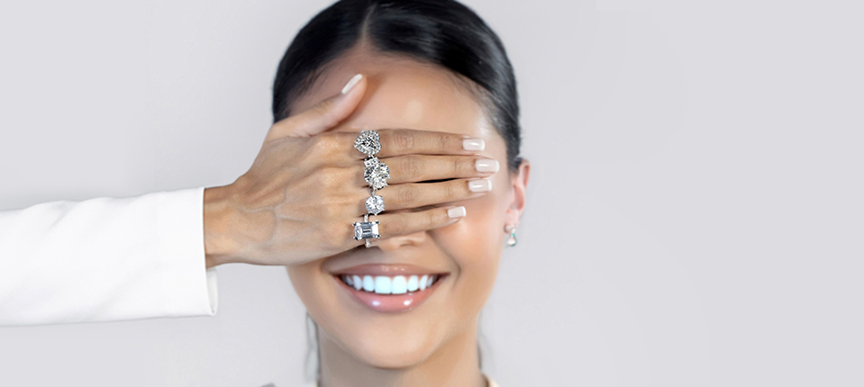Woman’s hand with elegant rings covers another woman’s eyes, smiling, illustrating various engagement ring styles.