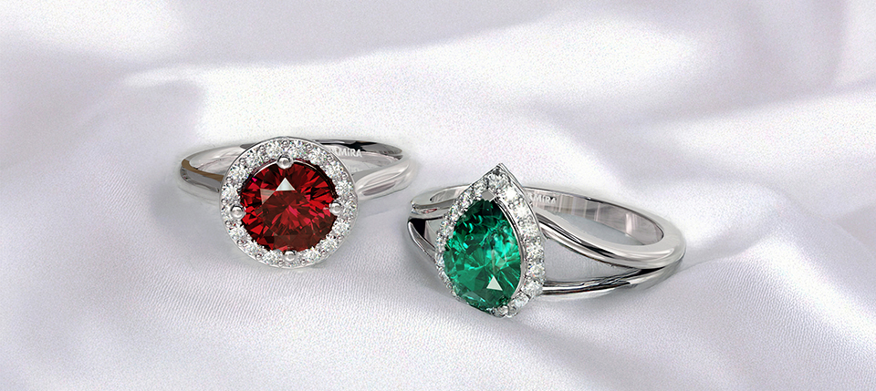 Two elegant engagement rings on white fabric, one with a red round gemstone and the other with a green teardrop gemstone, both accented with diamonds.