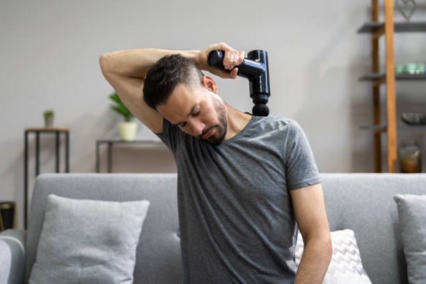 Man using a massage gun on his shoulder, ideal gift for boxers to relieve muscle tension, seated on a grey sofa at home.